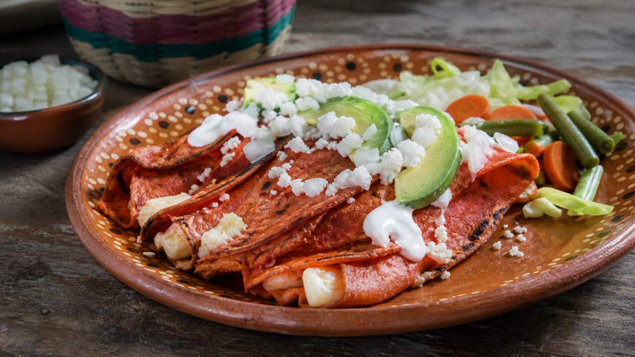 Enchiladas potosinas de masa rojiza con chile, queso, crema, aguacate y vegetales en plato de barro